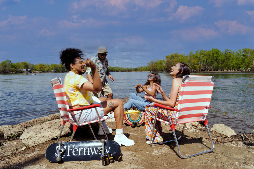 Four friends at the river's edge, laughing and enjoying a Pineapple Funk vape. A Fernway skateboard is leaning against one of the chairs they sit in.