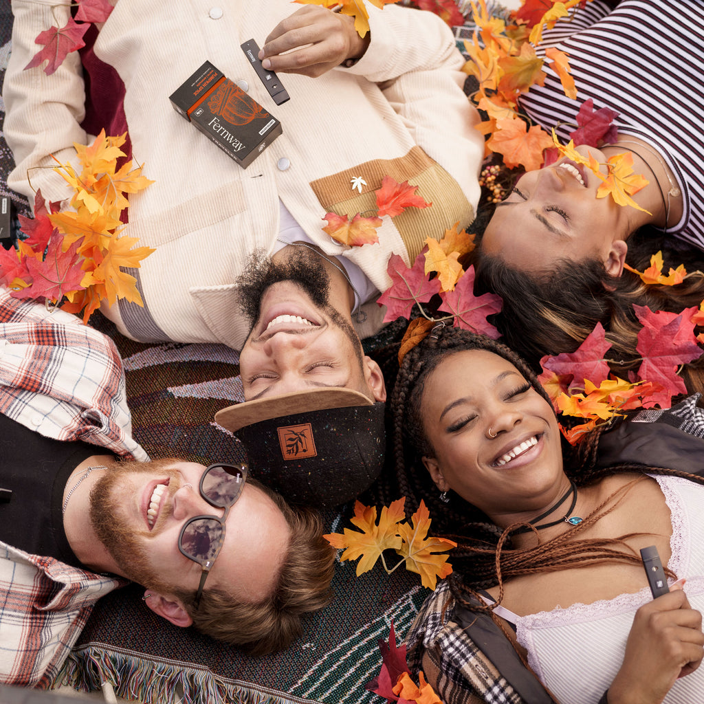 A group of friends lying on a blanket sharing a Pumpkin Spice vape and smiling at the falling autumn leaves.