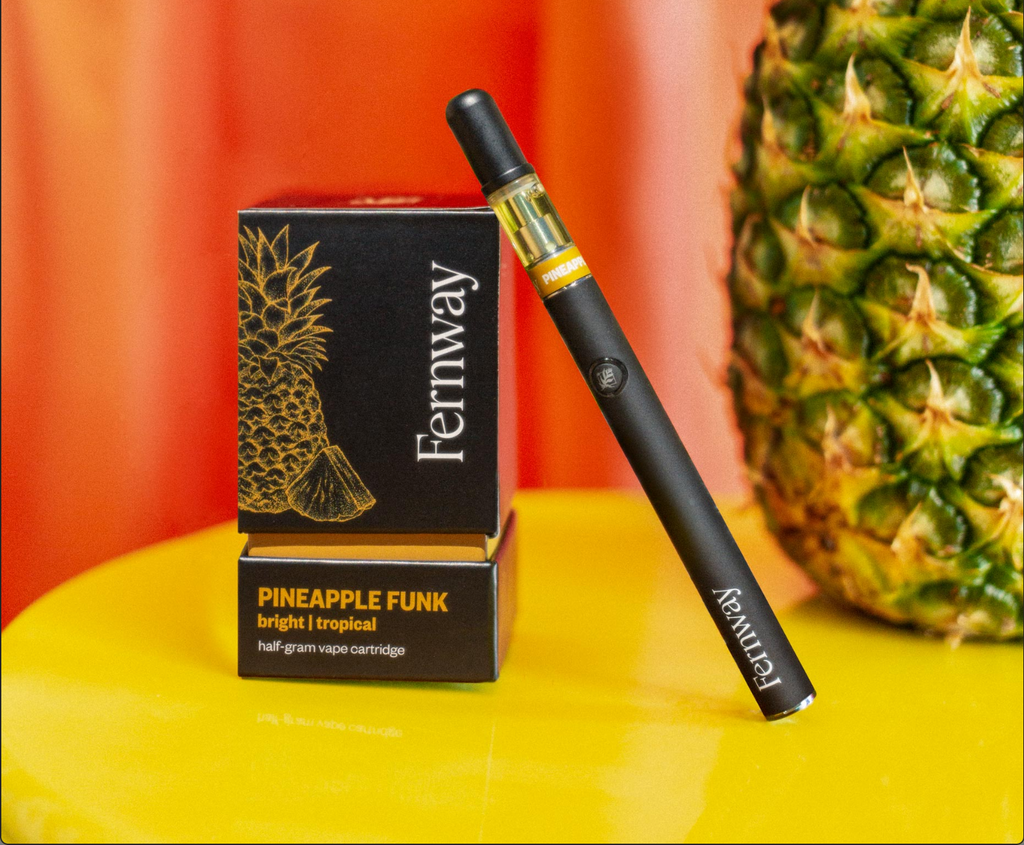A Pineapple Funk vape box with the pen leaning against it and an out of focus pineapple in the background.