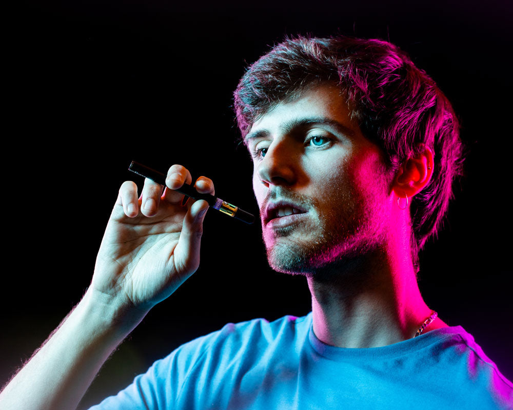 A young man with short hair and a blue shirt holding a Fernway Lavender Dream vape up to his mouth.
