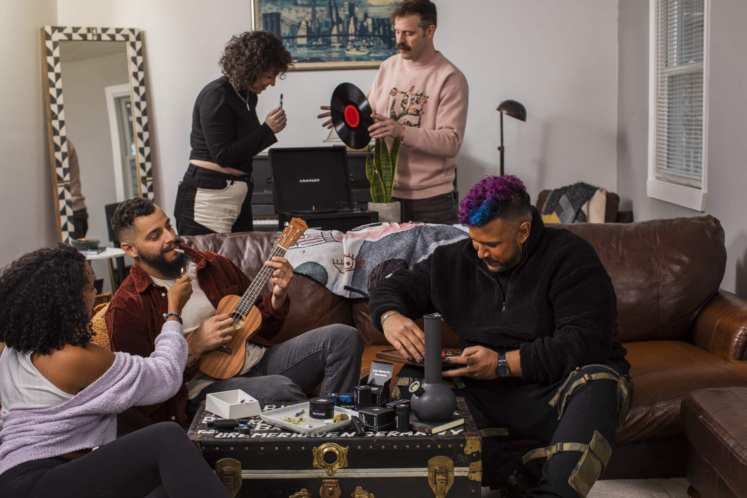 A group of friends - two women and three men - lounge around a living room smoking, playing music, and chilling.