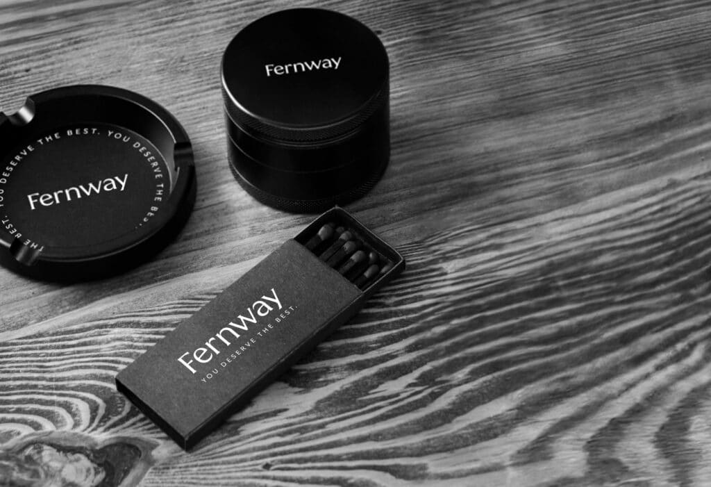 Black and white photo of several Fernway accessories - an ashtray, a grinder, and the box of matches.