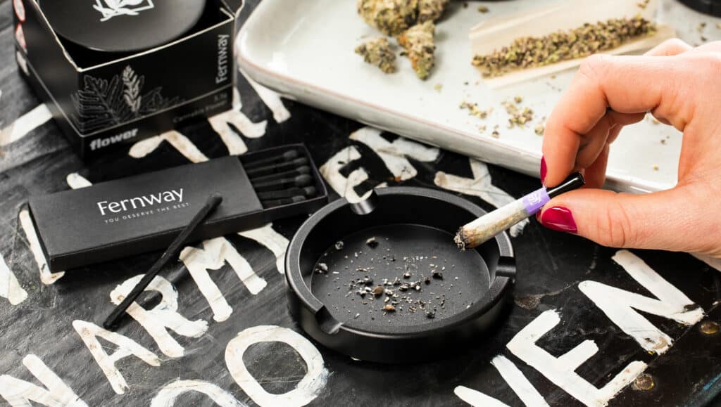 Photo of a hand ashing a Fernway joint into a Fernway ashtray, next to a Fernway matchbox, a Fernway Flower jar, and a rolling tray with some loose cannabis on it.